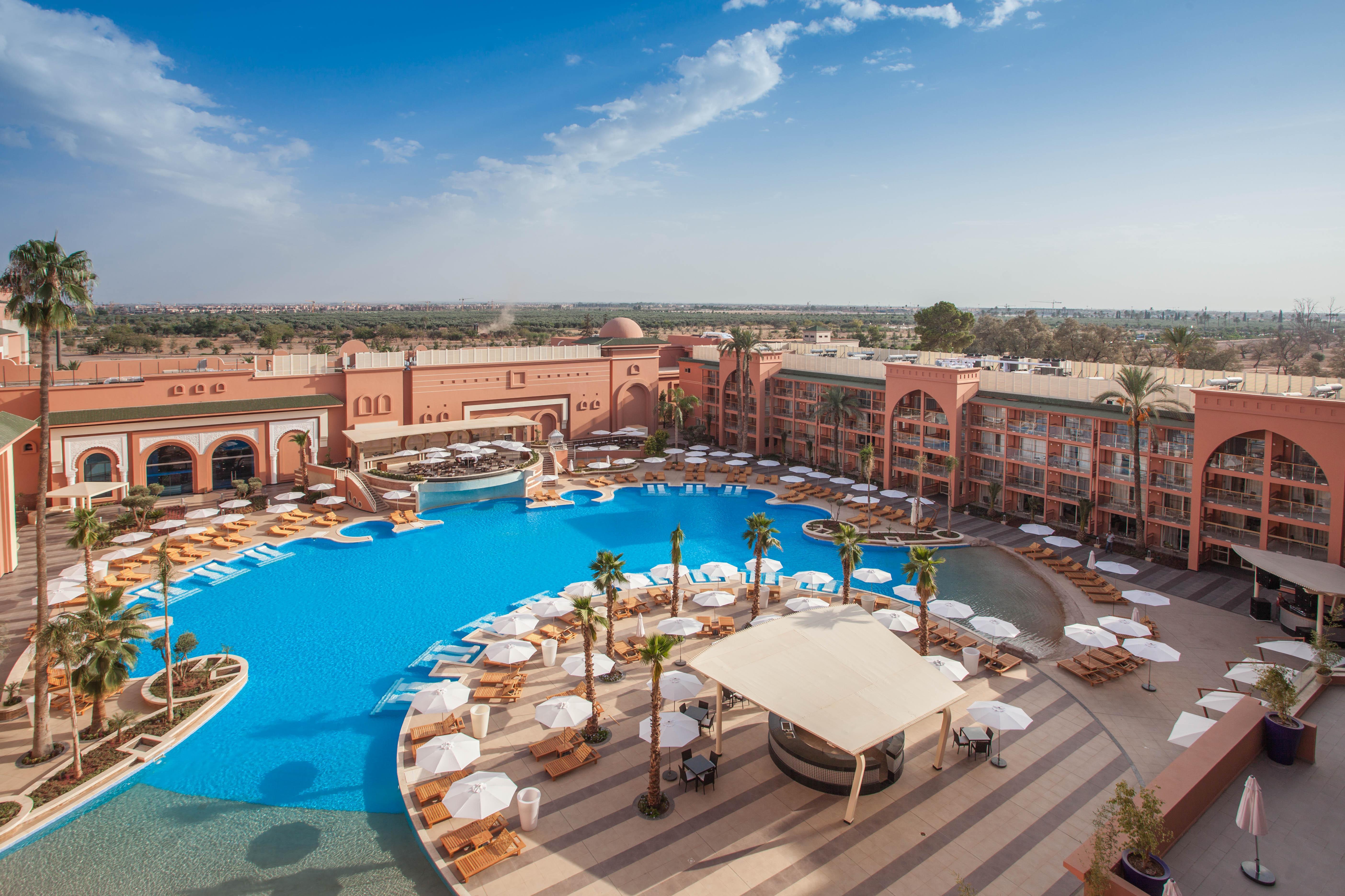 Savoy Le Grand Hotel Marrakech Marakeş Dış mekan fotoğraf The photo depicts a luxurious resort featuring a large, inviting swimming pool surrounded by sun loungers and umbrellas. The pool area is designed with a circular layout, and there are palm trees adding a tropical feel. In the background, there are s