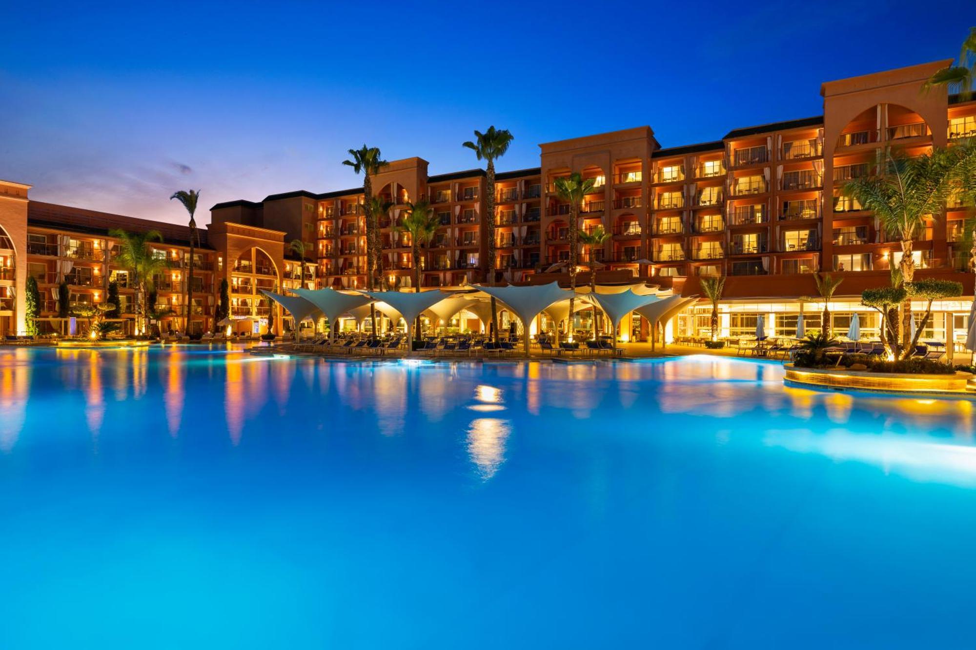 Savoy Le Grand Hotel Marrakech Marakeş Dış mekan fotoğraf The photo depicts a luxurious resort setting during twilight. There is a large, illuminated swimming pool in the foreground with a vibrant blue hue, reflecting the evening sky. Surrounding the pool are several palm trees, adding a tropical feel to th