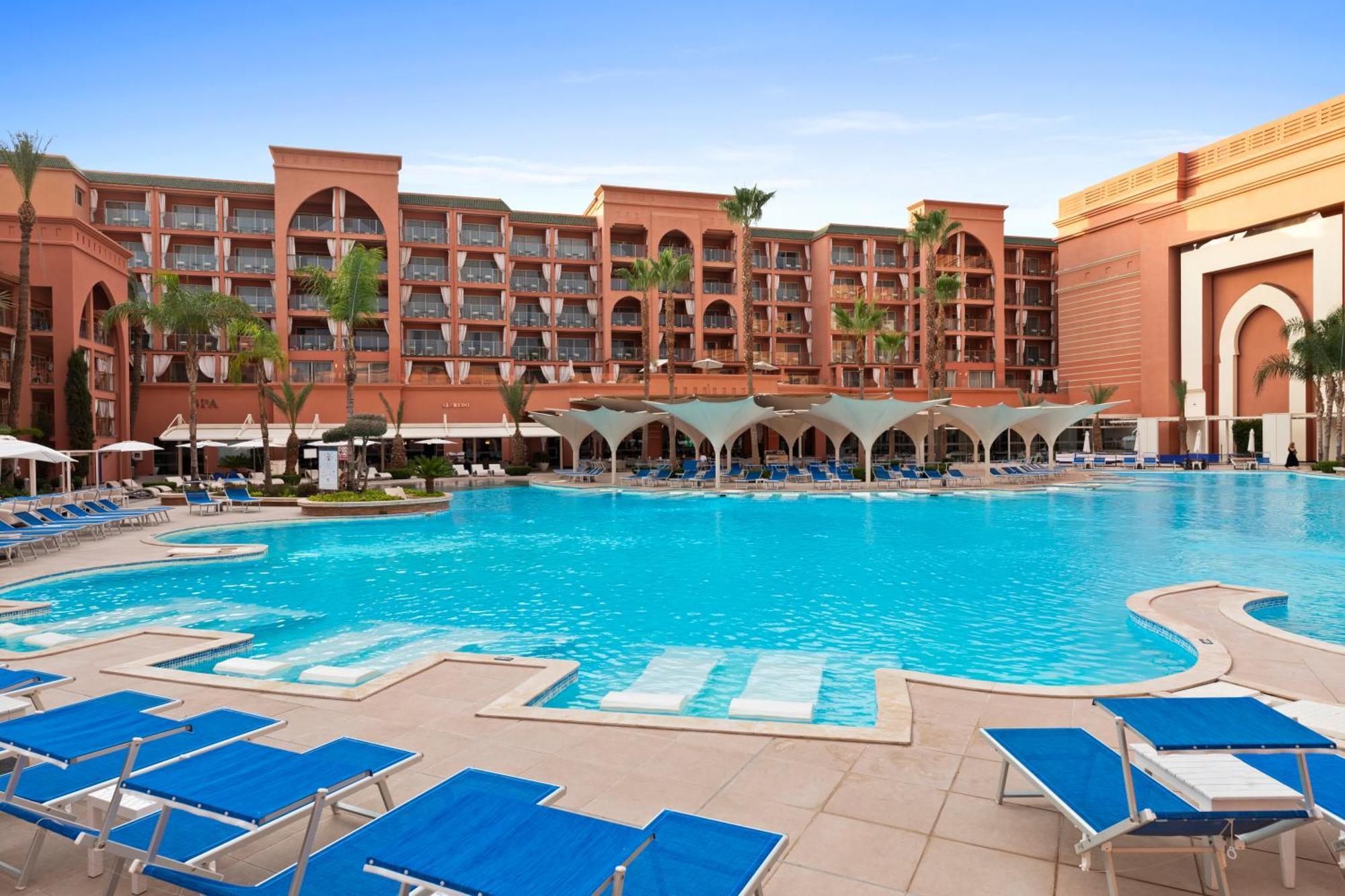 Savoy Le Grand Hotel Marrakech Marakeş Dış mekan fotoğraf The image shows a luxurious hotel setting featuring a large swimming pool in the foreground. The pool is surrounded by lounge chairs, primarily in blue, and has several shaded areas underneath canopies. In the background, there is a multi-story hotel