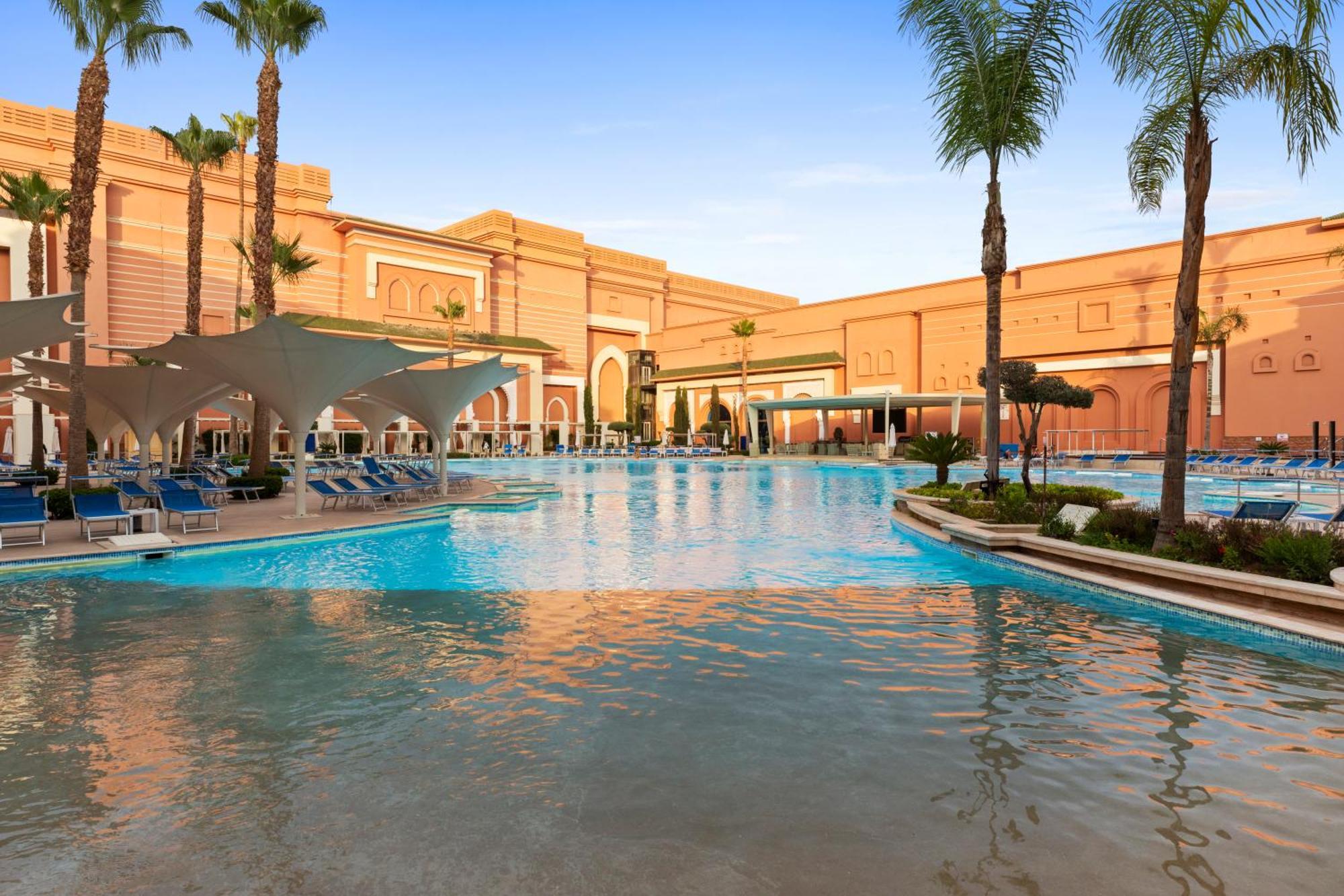 Savoy Le Grand Hotel Marrakech Marakeş Dış mekan fotoğraf The photo depicts a luxurious pool area surrounded by palm trees and a beautiful architectural backdrop. The water is clear and inviting, with lounge chairs neatly arranged along the poolside. The design features a blend of modern and classical eleme