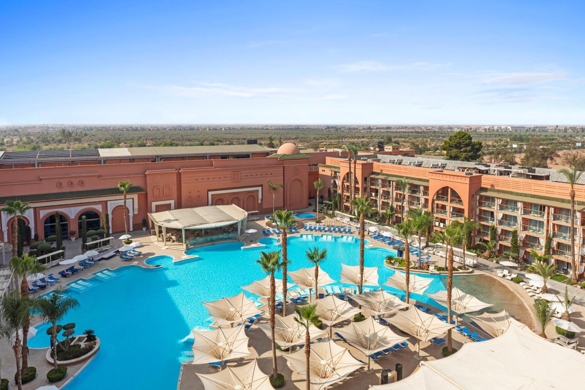 Savoy Le Grand Hotel Marrakech Marakeş Dış mekan fotoğraf The photo depicts a stylish resort pool area surrounded by palm trees. It features a large, inviting swimming pool with a spacious deck and lounge chairs arranged around it. There are shaded cabanas or umbrellas providing sun protection for guests. I