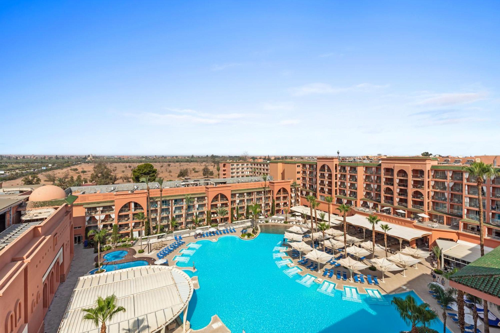 Savoy Le Grand Hotel Marrakech Marakeş Dış mekan fotoğraf The image shows a resort with a large swimming pool in the foreground, surrounded by lounge chairs and umbrellas. The pool area is adorned with palm trees, contributing to a tropical vibe. In the background, there is a multi-story building featuring 