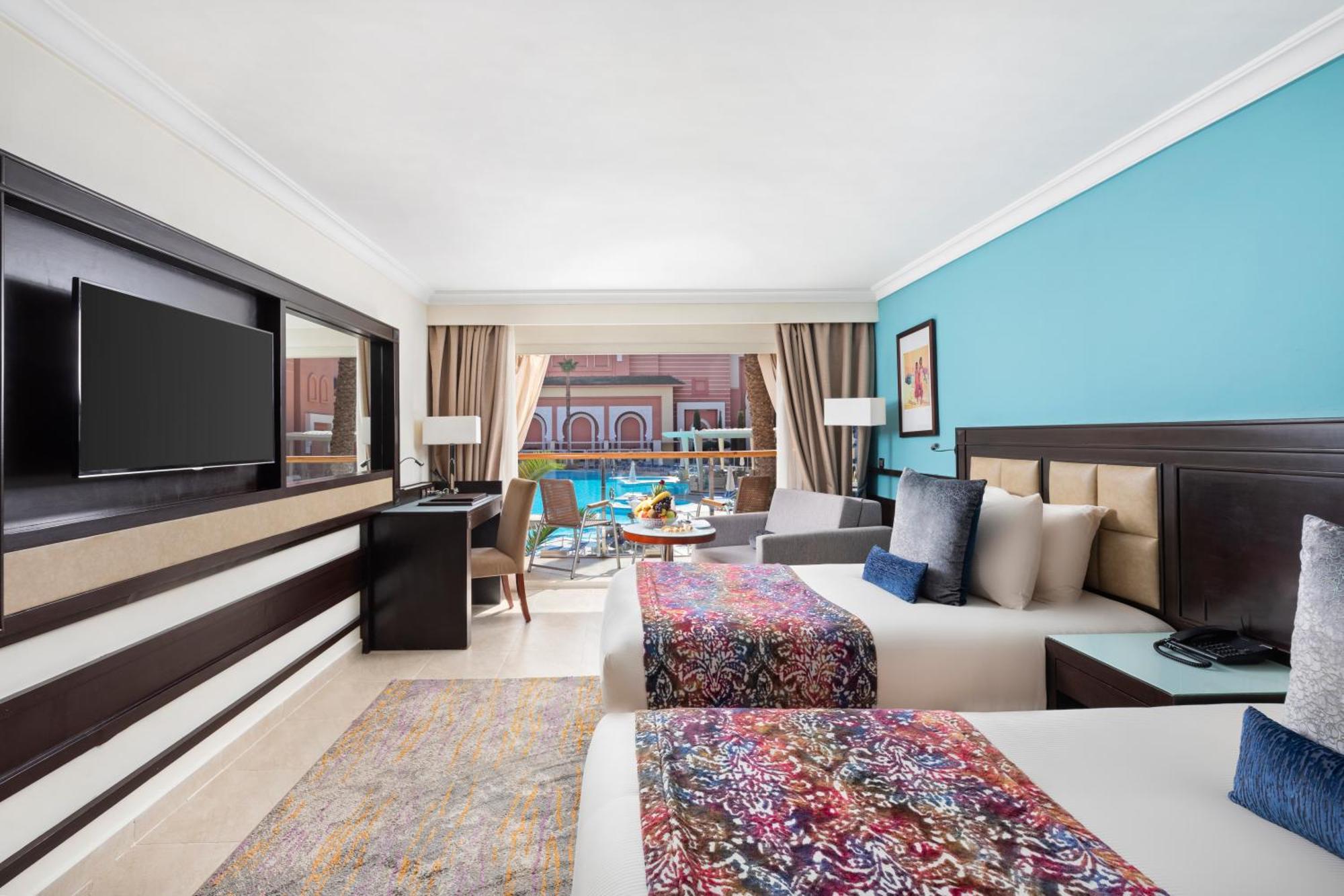 Savoy Le Grand Hotel Marrakech Marakeş Dış mekan fotoğraf The photo shows a stylish hotel room featuring two beds, each adorned with colorful patterned bedspreads. The walls are painted in a calming blue hue, and there are large windows allowing natural light to fill the space, with a view of an outdoor poo