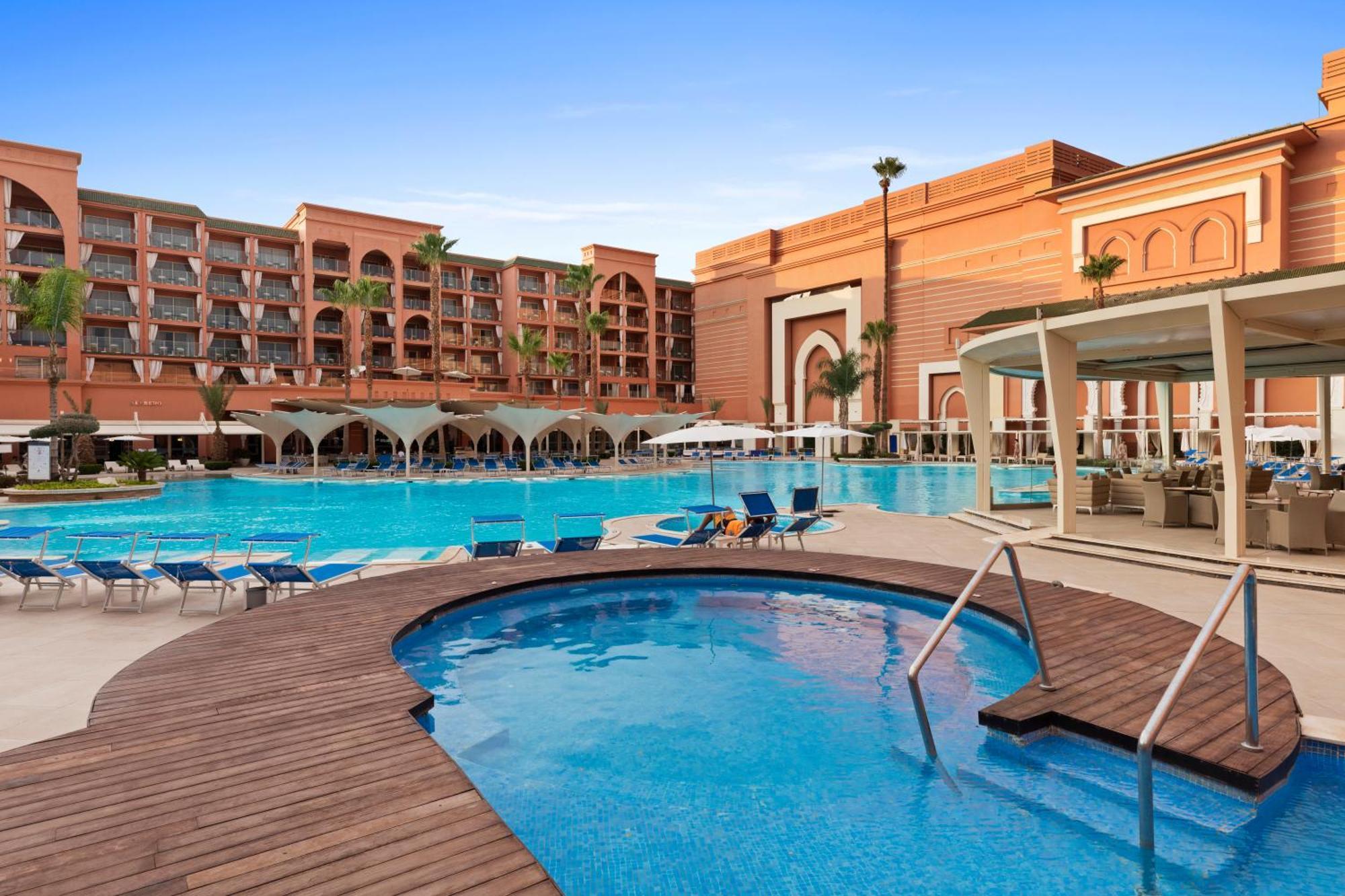 Savoy Le Grand Hotel Marrakech Marakeş Dış mekan fotoğraf The photo showcases a vibrant resort with a large swimming pool and a smaller jacuzzi area. The pool area is surrounded by lounge chairs, providing space for relaxation. In the background, there are several multi-story buildings designed with a Moroc