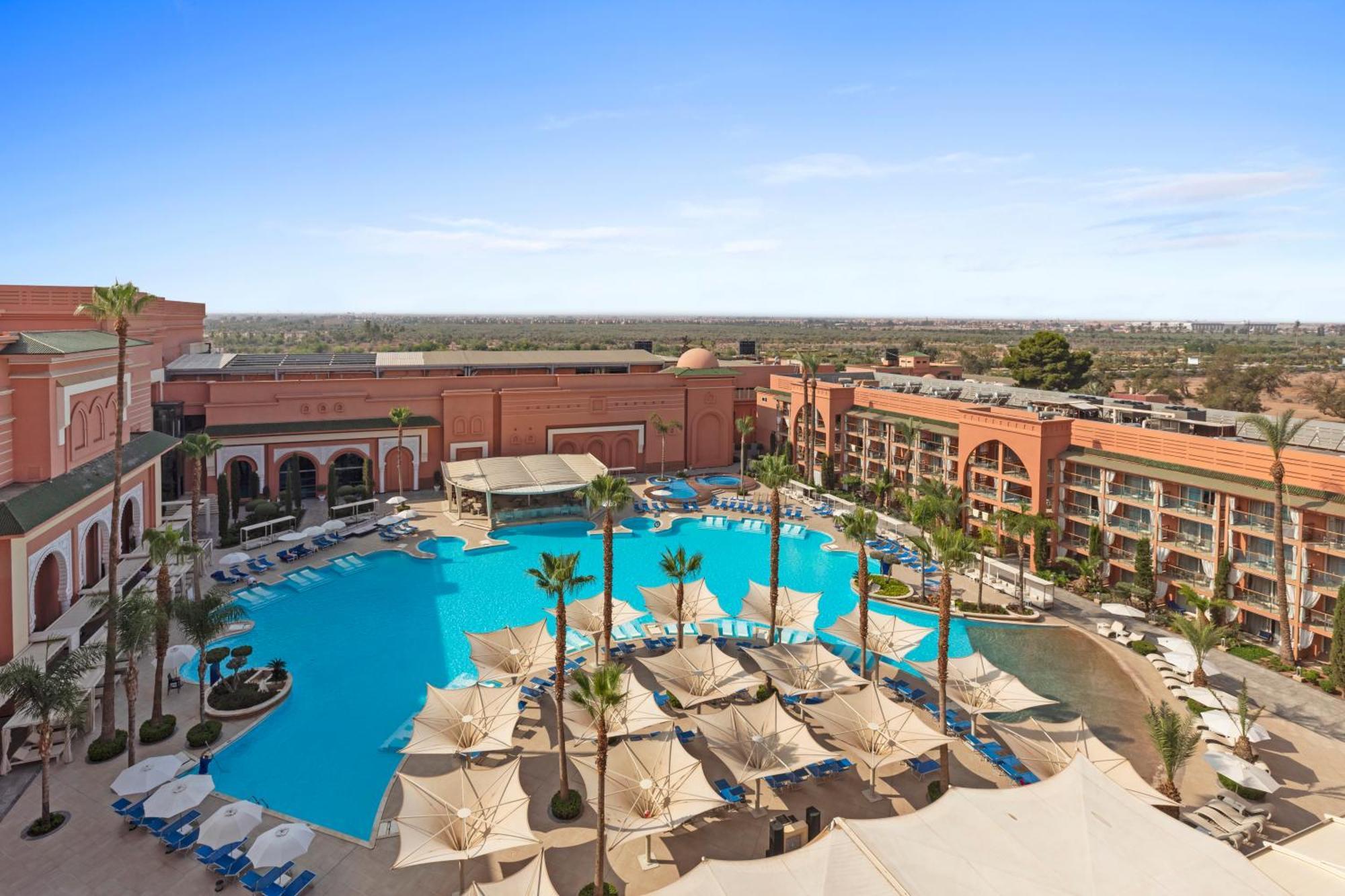 Savoy Le Grand Hotel Marrakech Marakeş Dış mekan fotoğraf The photo shows a luxurious hotel resort featuring a large swimming pool at the center. Surrounding the pool are several palm trees and sun loungers, suggesting a relaxing outdoor atmosphere. There are shaded cabanas or umbrellas providing cover for 