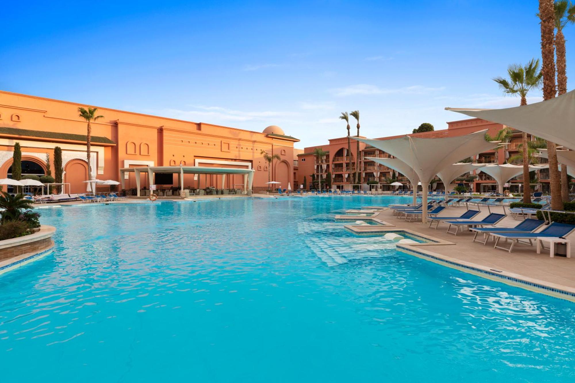 Savoy Le Grand Hotel Marrakech Marakeş Dış mekan fotoğraf The photo shows a large outdoor swimming pool with clear, turquoise water. Surrounding the pool are several lounge chairs, some of which are sheltered by large umbrellas. In the background, there are buildings with a distinctly warm-colored architect