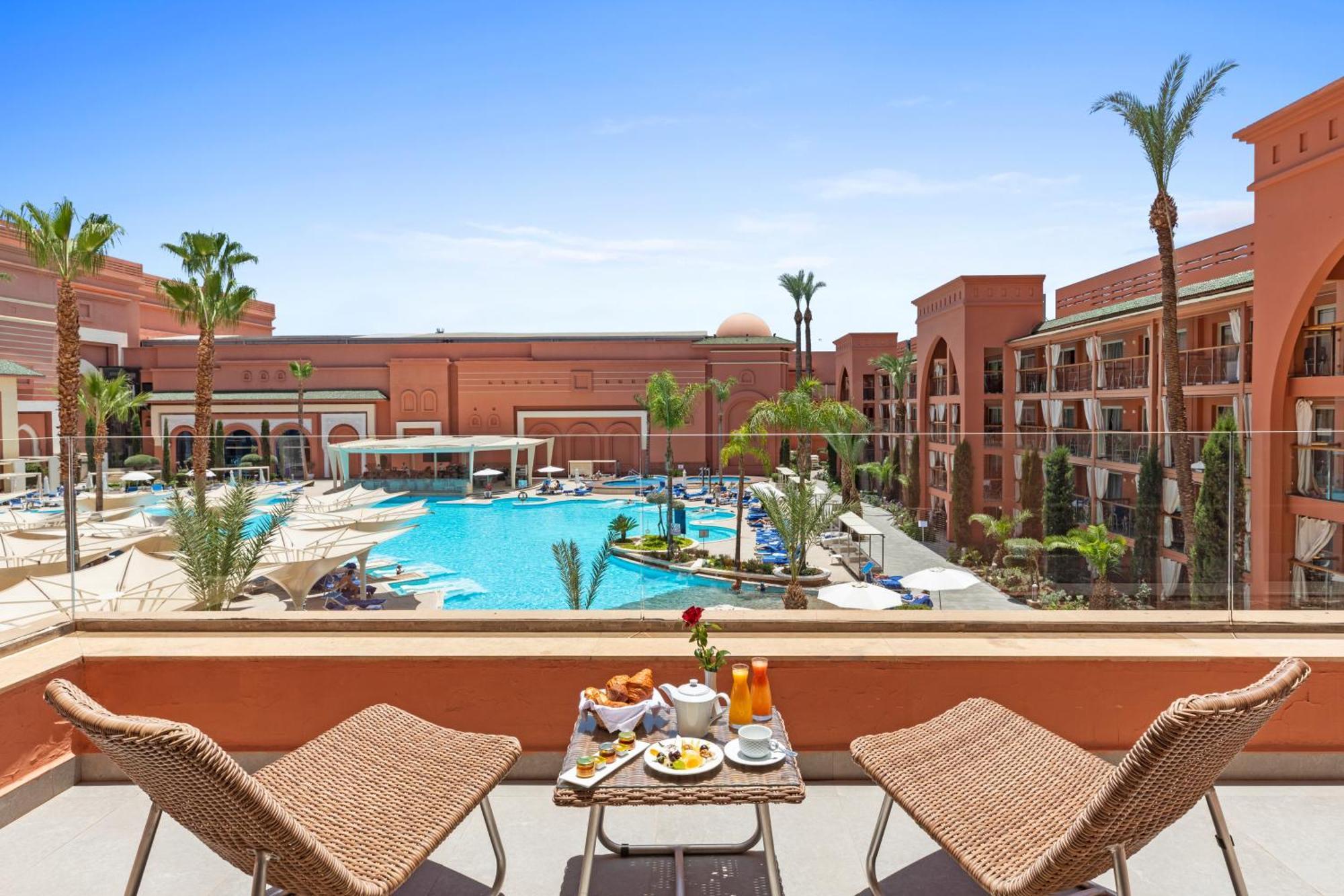 Savoy Le Grand Hotel Marrakech Marakeş Dış mekan fotoğraf The photo shows a picturesque view from a balcony overlooking a hotel pool area. In the foreground, there are two wicker chairs and a small table set with breakfast items, including a vase with a flower, a juice glass, and some food. In the backgroun