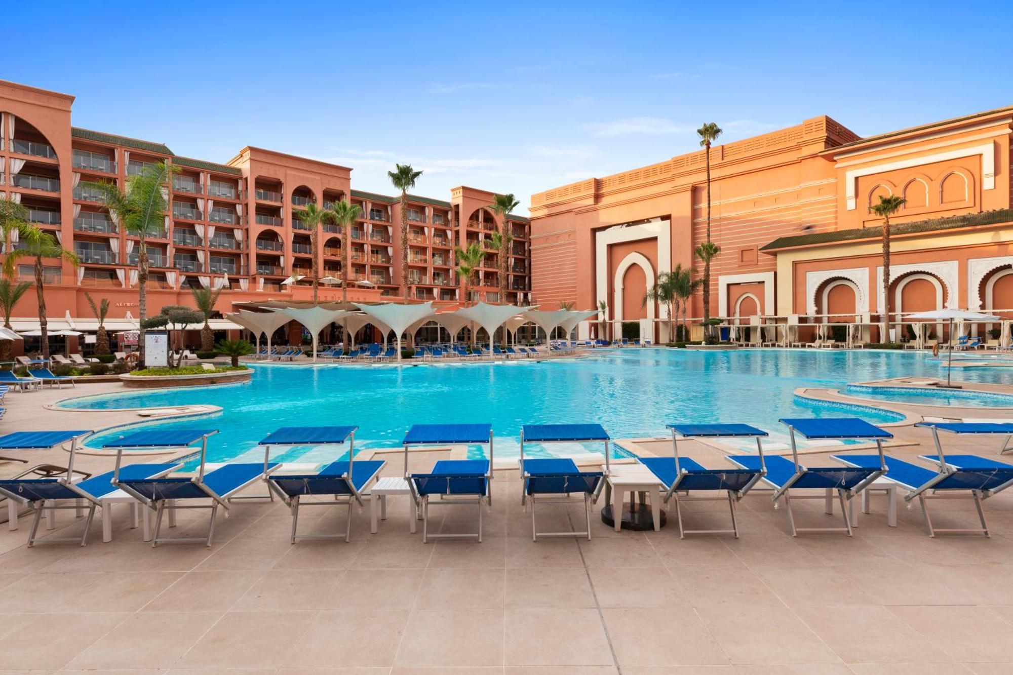 Savoy Le Grand Hotel Marrakech Marakeş Dış mekan fotoğraf The image shows a hotel or resort setting featuring a large swimming pool. The pool area is surrounded by lounge chairs that have blue cushions. In the background, there are several buildings with a desert-inspired architectural style, characterized 