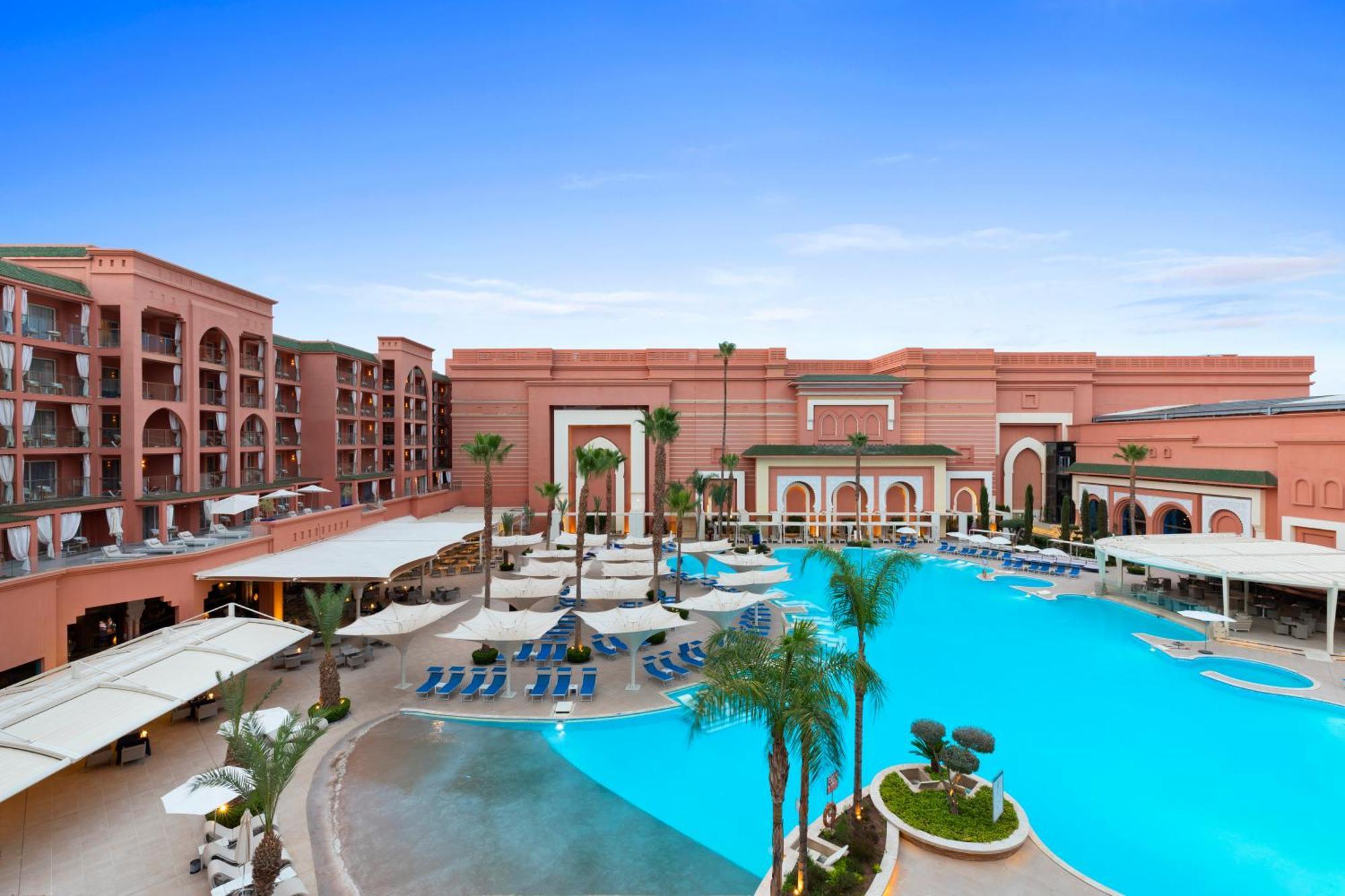 Savoy Le Grand Hotel Marrakech Marakeş Dış mekan fotoğraf The photo depicts a luxurious resort featuring an expansive pool area. The pool is a vibrant blue and surrounded by palm trees and lounge chairs. In the background, there are elegantly designed buildings with a reddish stone facade, characteristic of