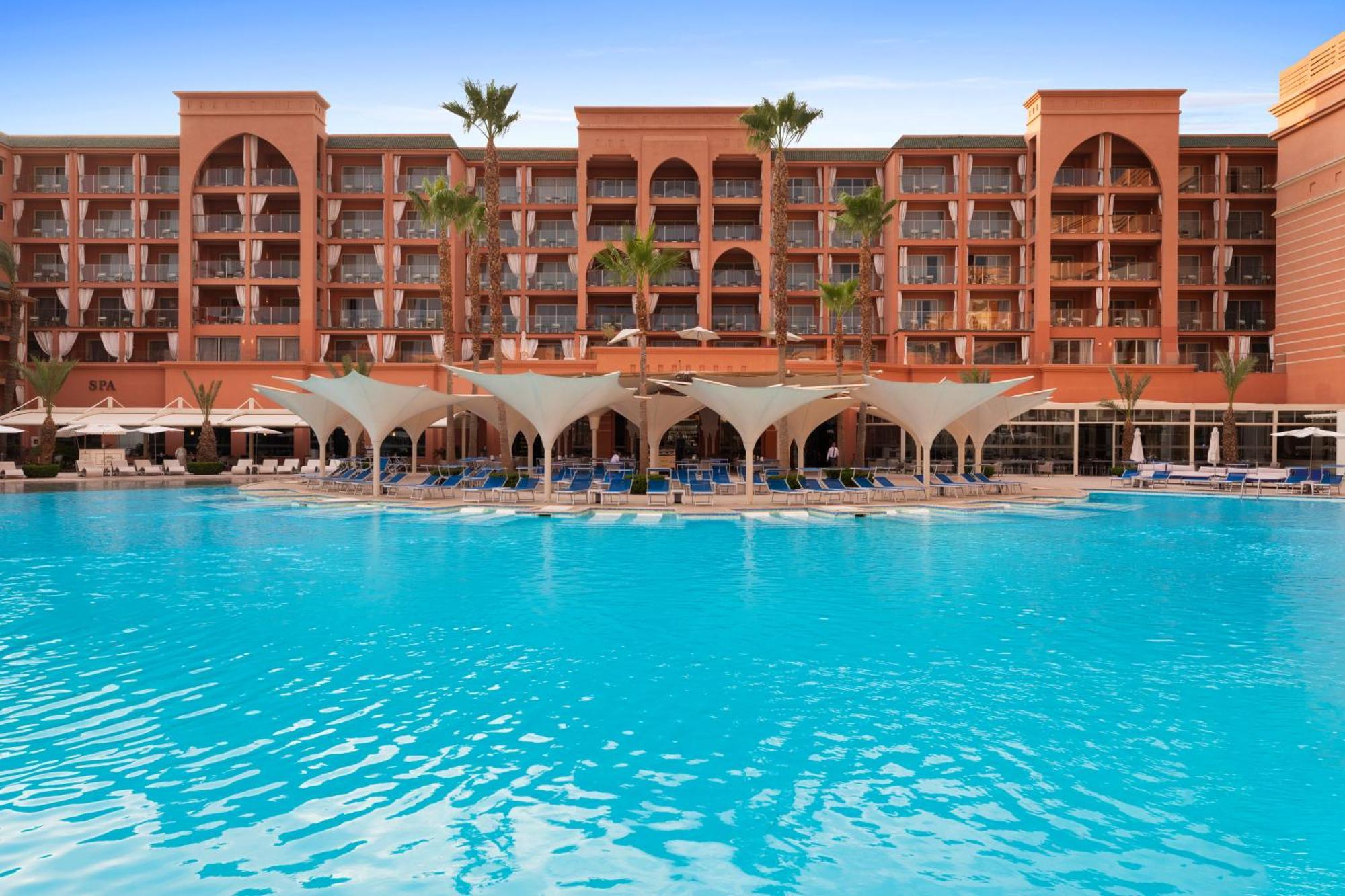 Savoy Le Grand Hotel Marrakech Marakeş Dış mekan fotoğraf The photo shows a beautiful hotel with an architectural style typical of Moroccan design, characterized by its red-orange facade and arched windows. In the foreground, there is a large swimming pool with crystal-clear blue water. The pool area is sha