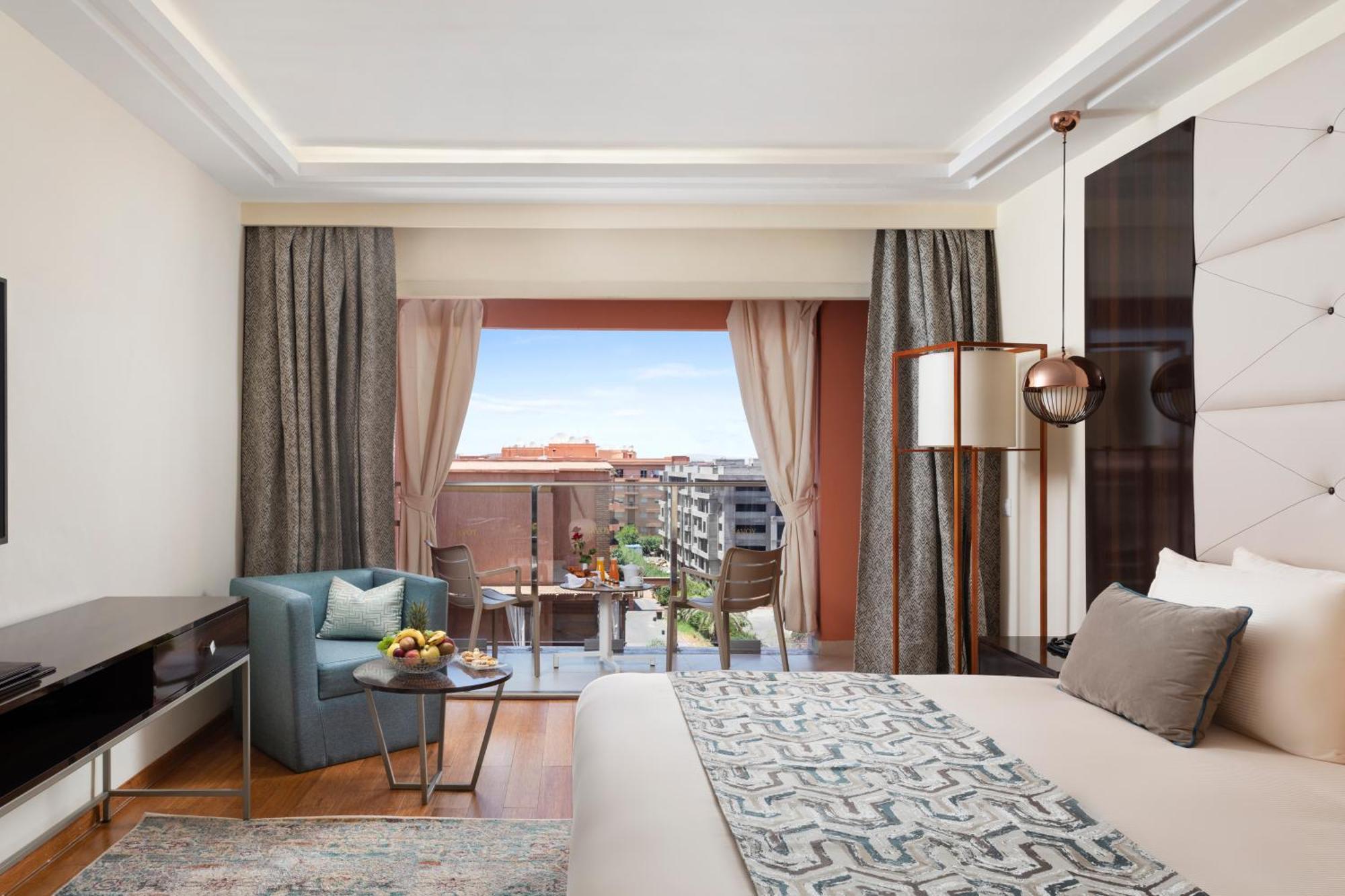 Savoy Le Grand Hotel Marrakech Marakeş Dış mekan fotoğraf The photo shows a modern hotel room featuring a comfortable bed with decorative pillows. There is a stylish area with a light blue armchair and a small table with fruit. The room has large windows with sheer curtains that reveal a scenic view outside