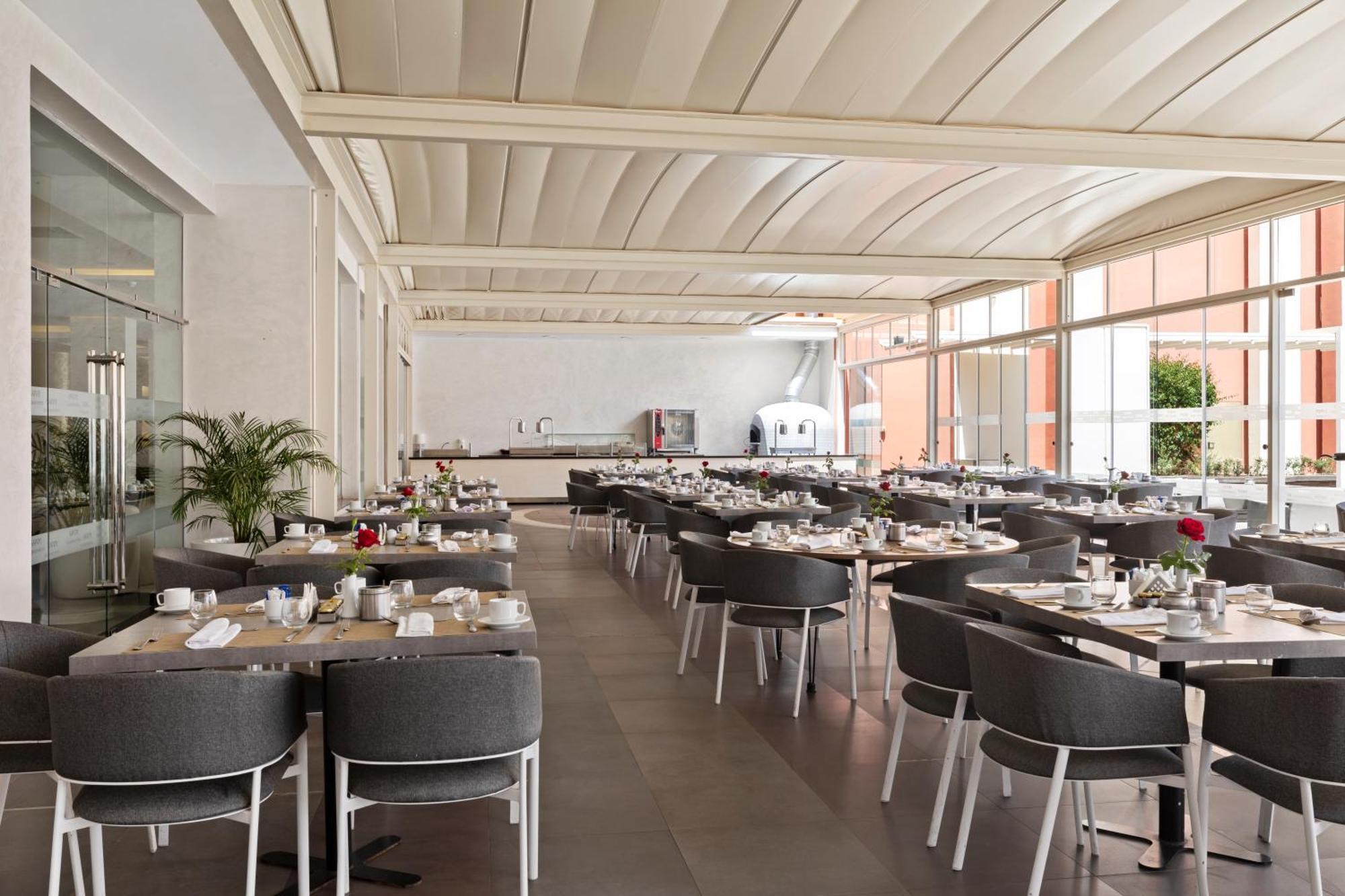 Savoy Le Grand Hotel Marrakech Marakeş Dış mekan fotoğraf The photo showcases a modern restaurant interior. The space features a bright, airy atmosphere with large windows allowing natural light to flood in. There are several tables set for dining, each with grey chairs and neatly arranged tableware includi