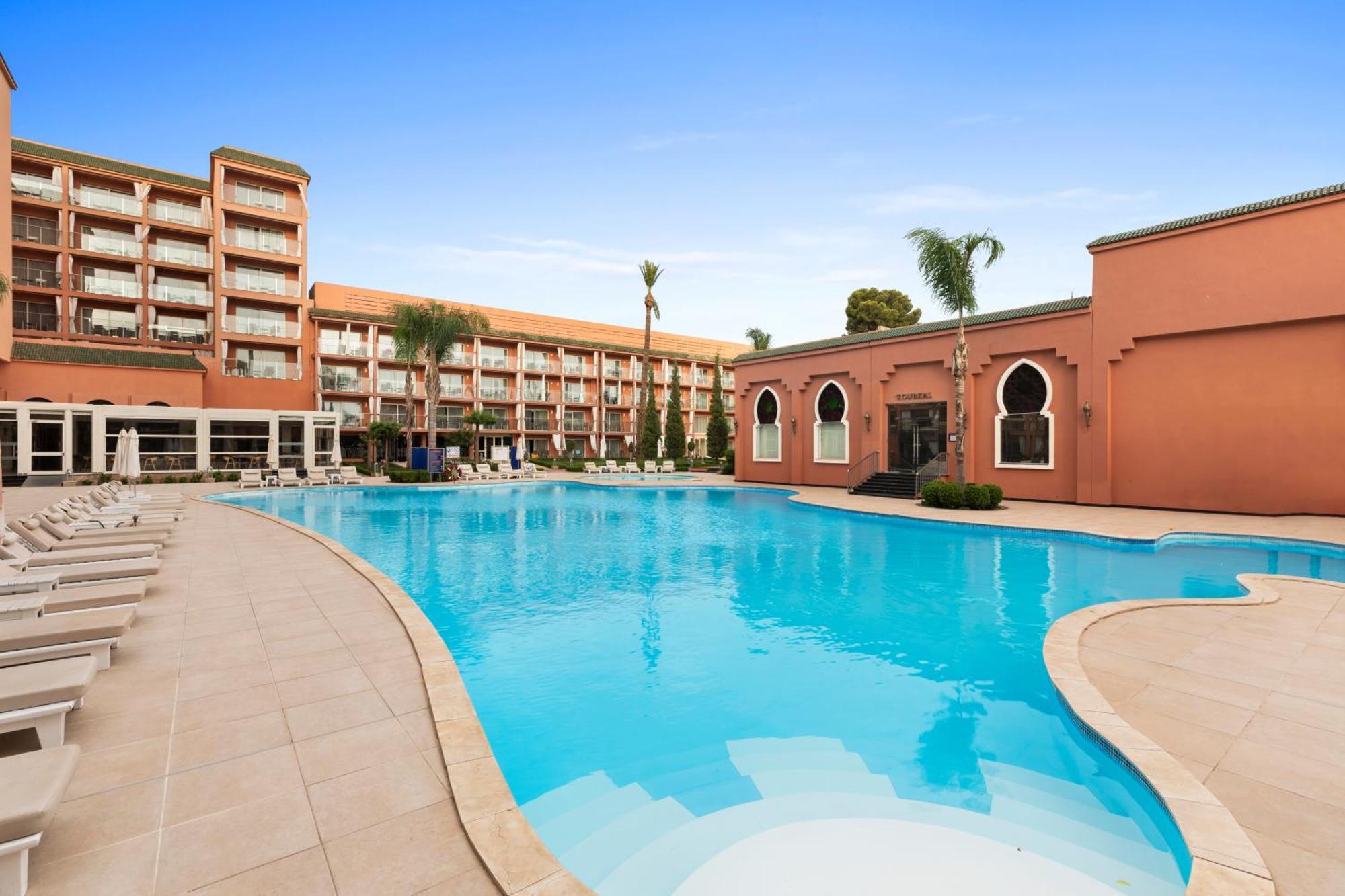 Savoy Le Grand Hotel Marrakech Marakeş Dış mekan fotoğraf The photo shows a large swimming pool in an outdoor setting, surrounded by palm trees. On one side of the pool, there is a modern building with multiple floors and balconies, while on the opposite side, there is a structure with a distinctive archite