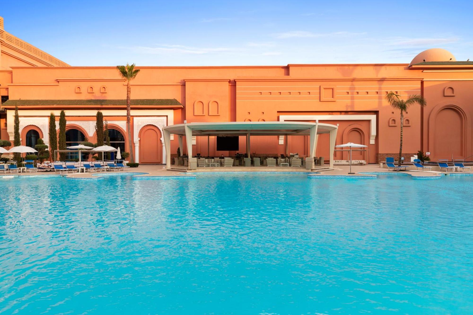 Savoy Le Grand Hotel Marrakech Marakeş Dış mekan fotoğraf The photo depicts a luxurious outdoor pool area of a resort or hotel. In the foreground, there is a large, inviting swimming pool with bright turquoise water. Surrounding the pool are comfortable lounge chairs and umbrellas, suggesting a relaxing atm