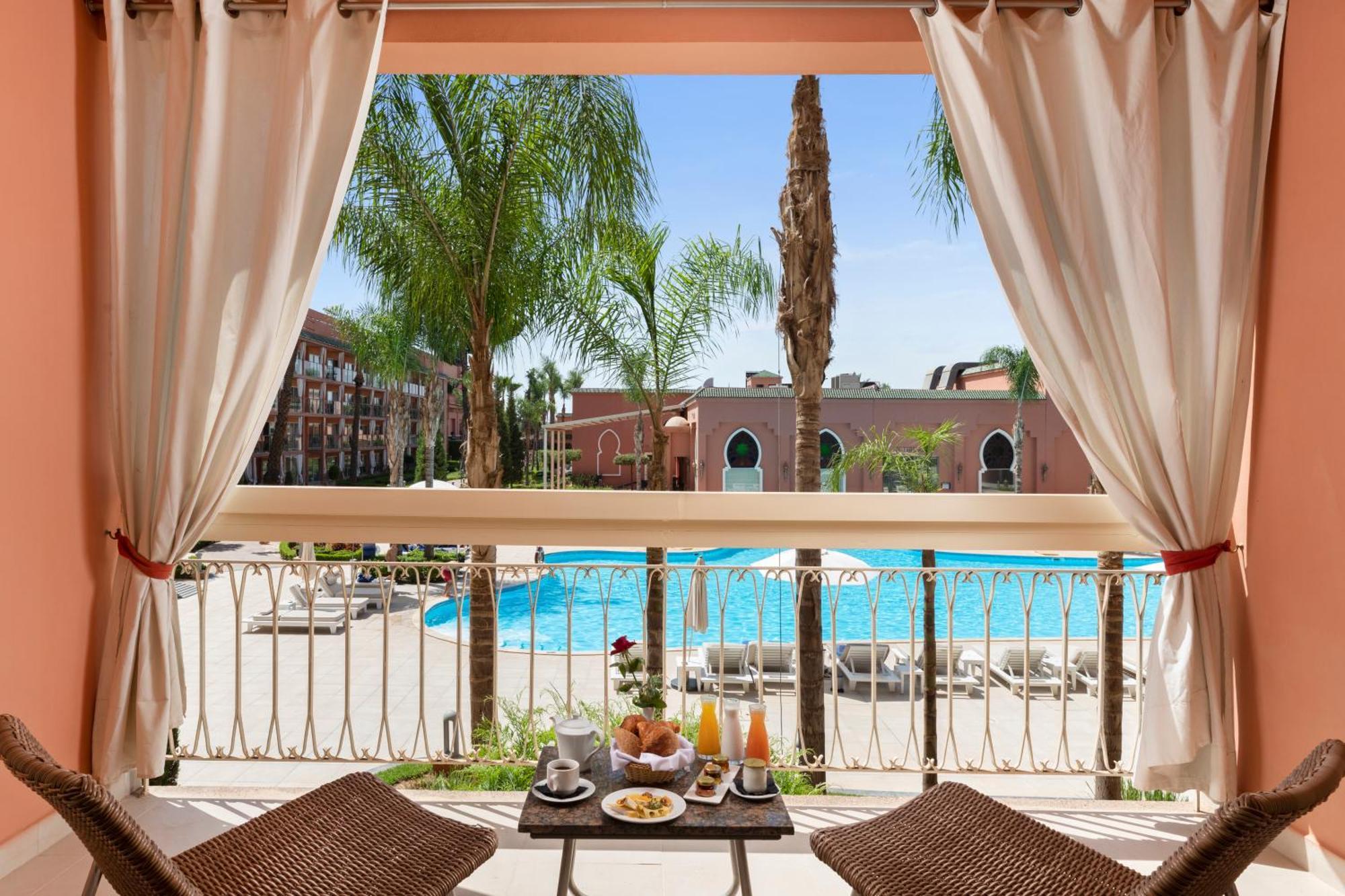 Savoy Le Grand Hotel Marrakech Marakeş Dış mekan fotoğraf The photo depicts a view from a balcony overlooking a swimming pool area. There are palm trees surrounding the pool, and several lounge chairs are arranged near the water. In the foreground, two wicker chairs are set up at a small table, which is ado