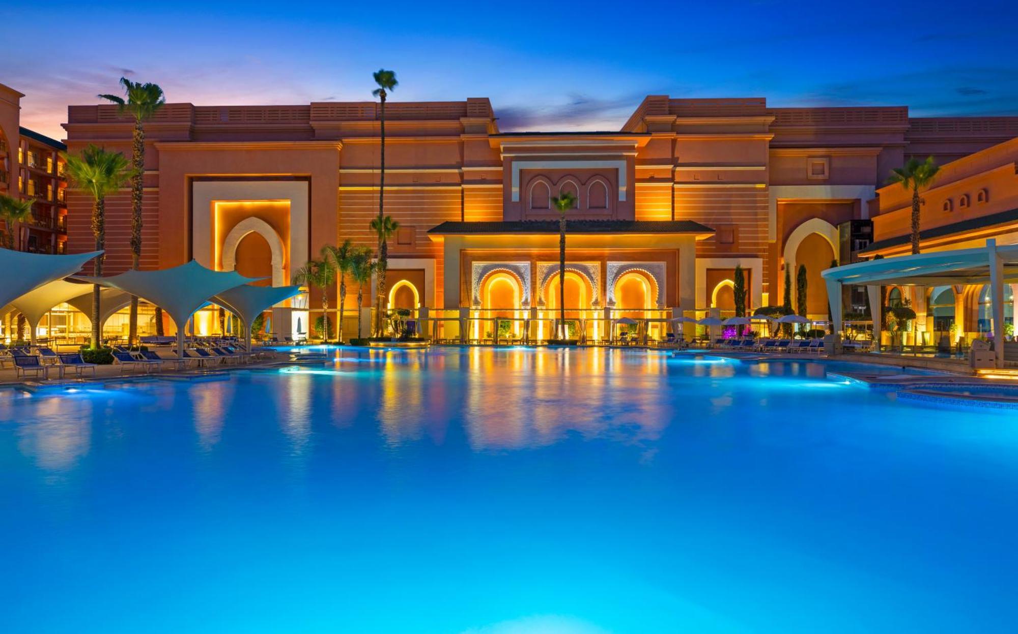 Savoy Le Grand Hotel Marrakech Marakeş Dış mekan fotoğraf The photo depicts a luxurious resort featuring a large, illuminated swimming pool in the foreground. The pool is surrounded by palm trees and lounge chairs, reflecting vibrant blue water. In the background, there is an elaborate building designed wit