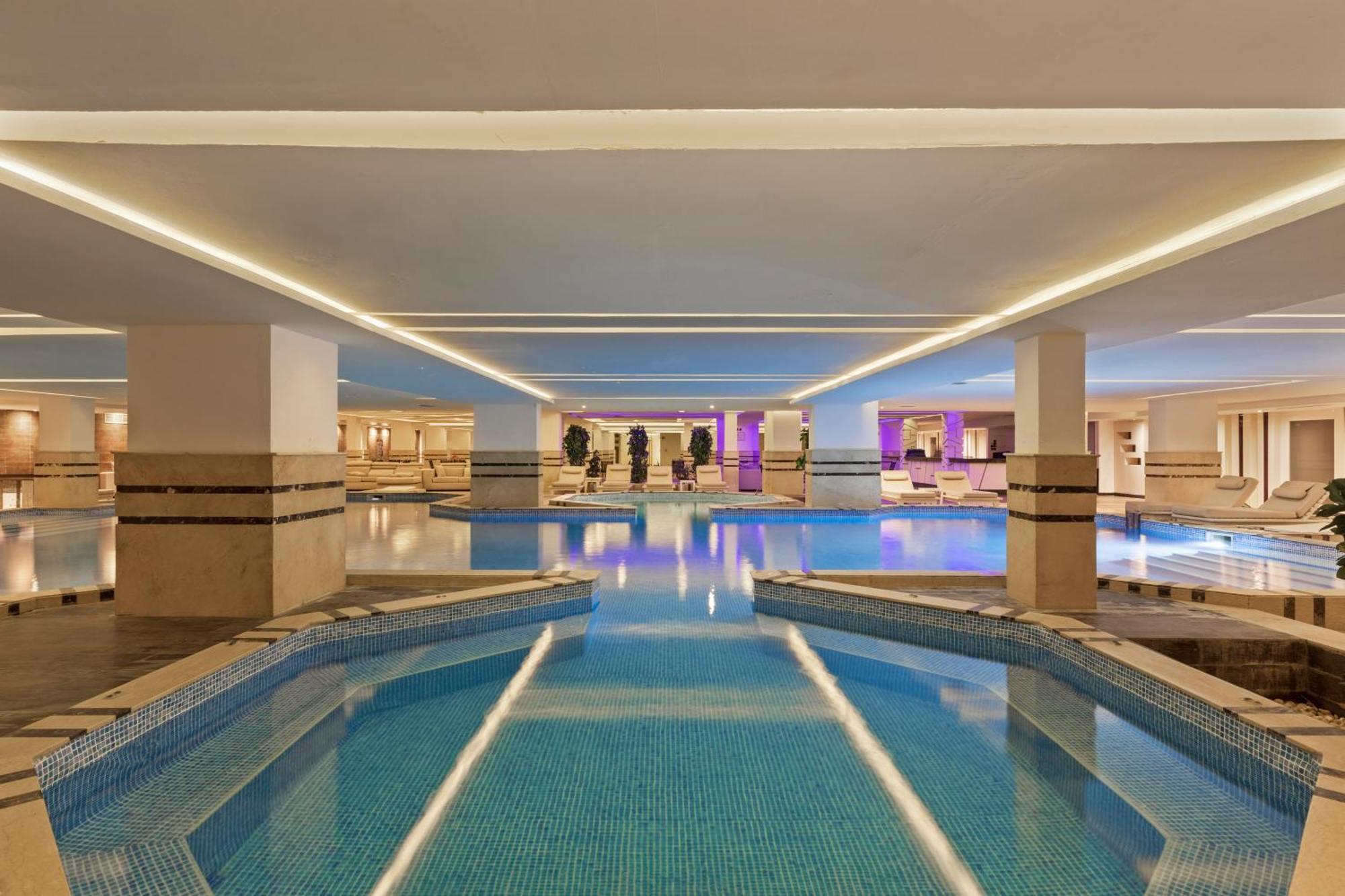 Savoy Le Grand Hotel Marrakech Marakeş Dış mekan fotoğraf The photo shows an interior swimming pool area that is well-lit with soft lighting. The pool features a unique shape that includes curved sections and pathways. The water is a bright blue and appears inviting. Surrounding the pool are columns and wal