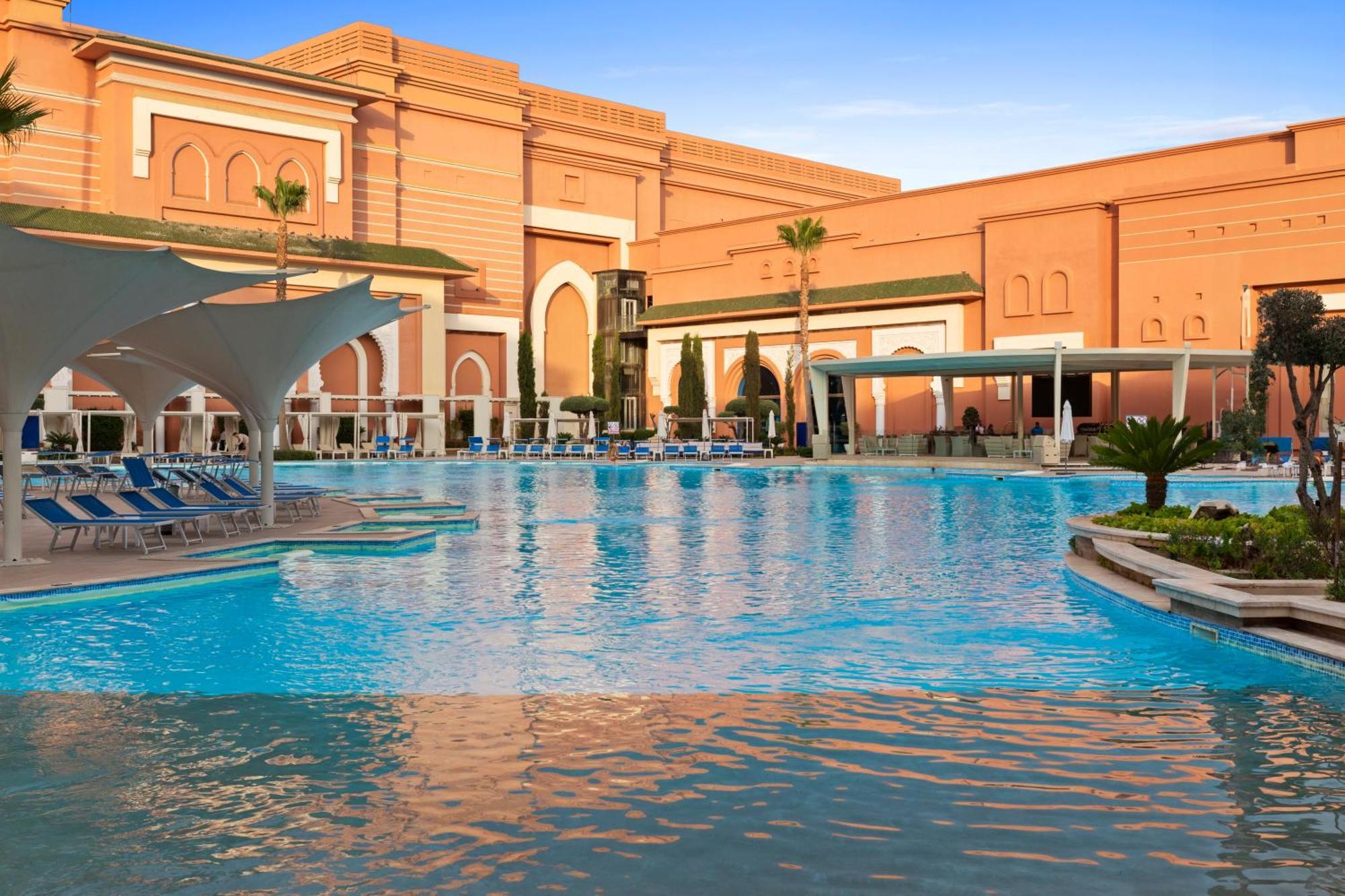 Savoy Le Grand Hotel Marrakech Marakeş Dış mekan fotoğraf The photo shows a large, inviting swimming pool with clear blue water, surrounded by lounge chairs. In the background, there is an elegantly designed building with a warm-colored facade, featuring arches and decorative elements, typical of luxury res