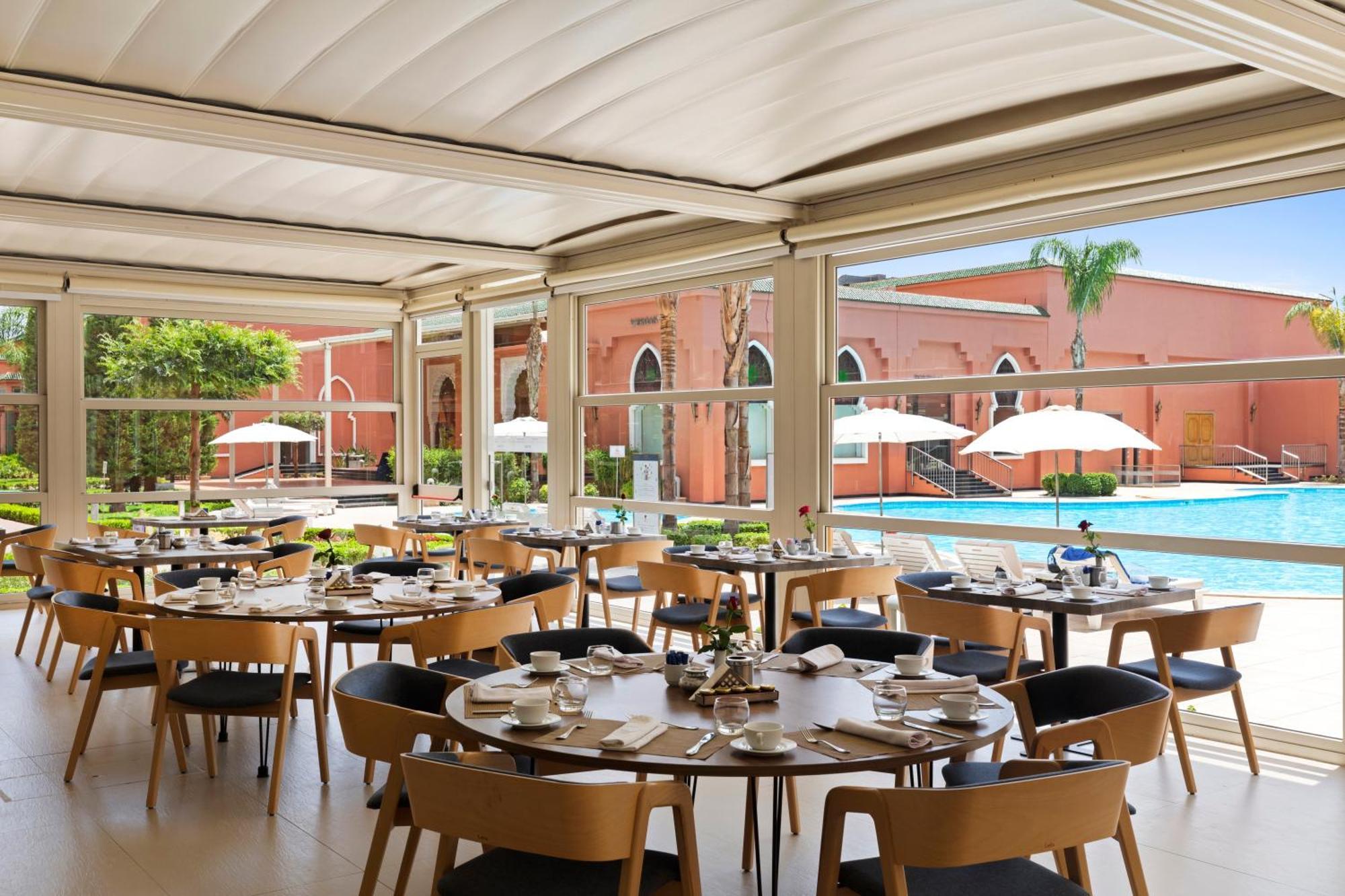 Savoy Le Grand Hotel Marrakech Marakeş Dış mekan fotoğraf The photo shows a modern restaurant setting with several round tables arranged for dining. Each table is neatly set with plates, glasses, and cutlery. The restaurant features large windows that allow plenty of natural light to flood in, offering a vi