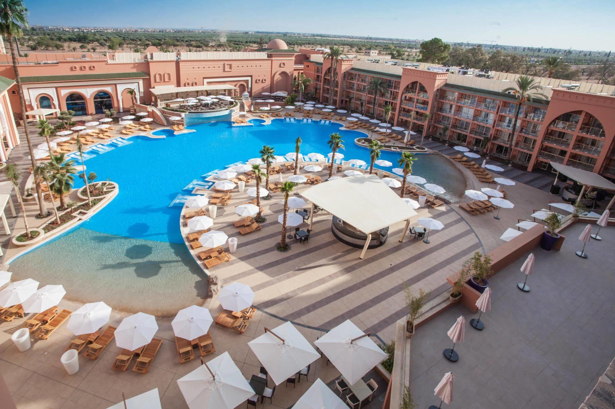 Savoy Le Grand Hotel Marrakech Marakeş Dış mekan fotoğraf The photo shows a large, luxurious hotel resort with a spacious swimming pool at its center. The pool area features several lounge chairs and umbrellas arranged around the water, providing a relaxing atmosphere for guests. Surrounding the pool are la